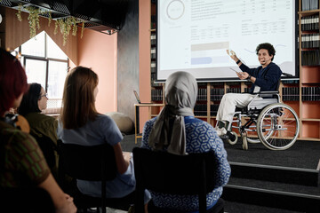 Cheerful Middle Eastern student with disability in wheelchair doing presentation at conference in...