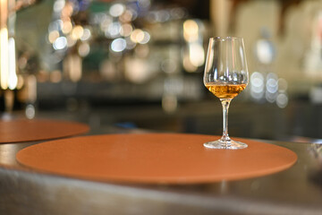 Single glass of white wine stands oncounter, with blurred background of bottles and bar atmosphere