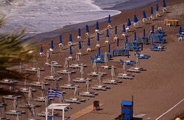 Sea coast with white waves and several rows of sun loungers and umbrellas on a sand and pebble beach. Next to the sunbeds there is a wooden blue changing room and a Greek flag.