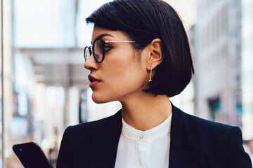 Serious female trader in eyewear walking on street of megalopolis with tasty coffee in...