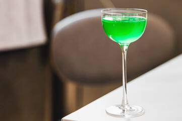 A vibrant green cocktail in a classic stem glass on a white surface with a blurred background.