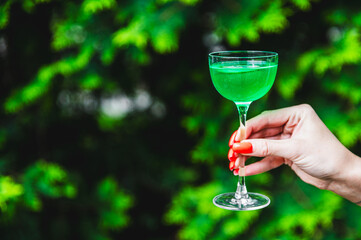 A close-up of a hand holding a glass with a green beverage against a blurred background of lush...