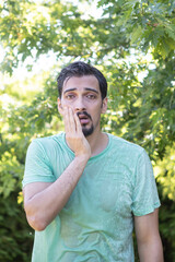 A wet and upset man stands in the park. The guy was caught in a sudden rain in broad daylight	