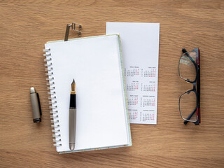 detail seen from above of a work table. Calendar, notebook open to a blank page, fountain pen,...