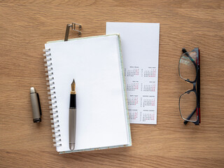 detail seen from above of a work table. Calendar, notebook open to a blank page, fountain pen, bookmark and glasses on a wooden table