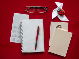 Overhead view of a desk. Red table and objects arranged in an orderly manner. Notebooks, pencils, glasses, calendar, a paper bow tie and a stone paperweight painted by a child