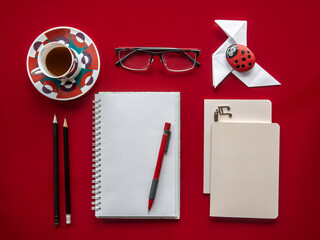 Overhead view of a desk. Red table and objects arranged in an orderly manner. Notebooks, pencils, glasses, cup of coffee, a paper bow tie and a stone paperweight painted by a child