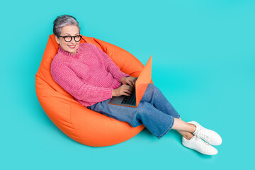 Full length photo of smart successful woman wear pink sweater lay on bean bag typing message on laptop isolated on teal color background