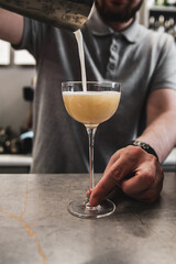 A person pours a creamy beverage into a stemmed glass on a bar counter, capturing dynamic motion...