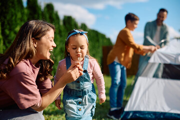 Small girl blowing dandelion with her mother during family camping.