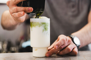 Fototapeta na wymiar Hands pour green liquid into a white creamy beverage, likely a matcha latte, on a marble countertop