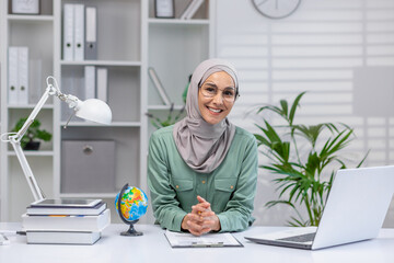 Portrait of happy teacher, woman in hijab sitting at desk smiling and looking at camera, muslim...