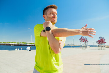 A man in a bright yellow shirt stretches his arm outdoors on a sunny day by a waterfront.