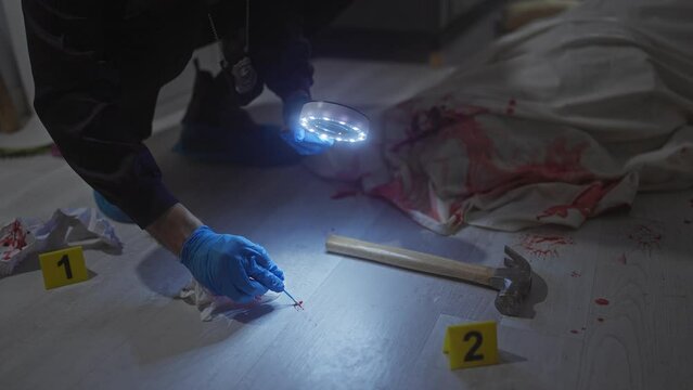 An investigator examines evidence at a night-time indoor crime scene with a body, hammer, and bloodstains