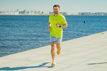 A man in a yellow shirt and shorts runs along a sunny waterfront promenade, smiling, with the city...