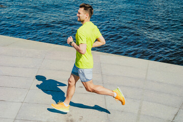 A man in a yellow shirt and shorts runs along a sunny waterfront promenade, with the blue water...