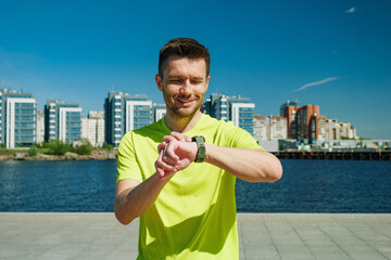 A man in a yellow shirt checks his wristwatch while standing on a sunny waterfront promenade, with...