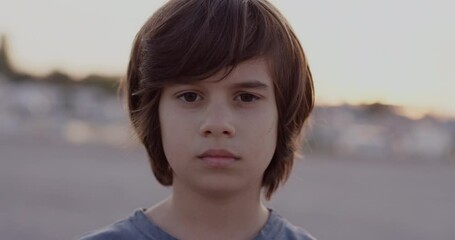 A young boy with long hair stands outdoors, looking directly at the camera with a serious expression on his face.