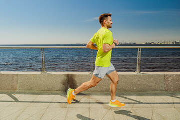 A man in a yellow shirt and shorts jogs along a sunny waterfront promenade, with blue water and a...