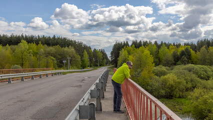 A solitary man in a yellow jacket and sunglasses stands on a bridge, gazing over the railing at a...