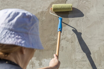 A woman in a blue vest, blue shirt and hat primes a textured concrete wall with a roller and brush....