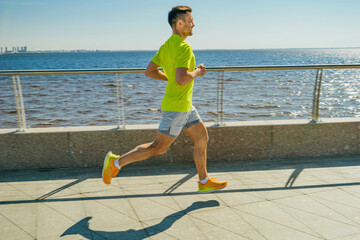 A man in a yellow shirt and shorts jogs along a sunny waterfront promenade, with blue water and...