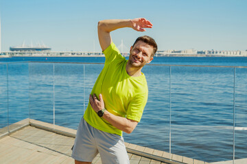 A man in a yellow shirt performs a side stretch by the waterfront, with a clear blue sky and...