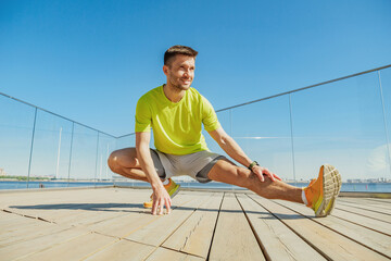 A man in a bright yellow shirt performs a deep side lunge stretch on a wooden deck, overlooking a...