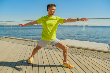 A man in a bright yellow shirt performs a wide-leg stretch with arms extended on a wooden deck by...