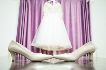 A bridal setting. In the foreground, there are two high-heeled shoes, one in a light beige color and the other in a slightly darker shade. Behind these shoes, a white wedding gown