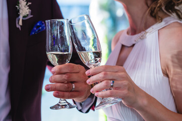 They are holding champagne glasses, clinking them together. The man is wearing a dark suit with a blue boutonnière on his lapel, while the woman dons a white dress.