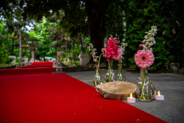 An outdoor setting with a vibrant red carpet leading the viewer's eye towards a group of tall trees in the background. On the carpet, there are three clear glass vases containing pink and white flower