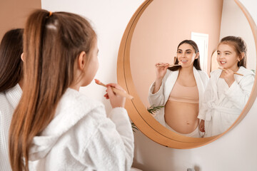 Little girl with her pregnant mother brushing teeth near mirror in bathroom