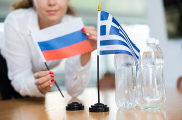 Young female assistant setting up greek and russian flags for international negotiations