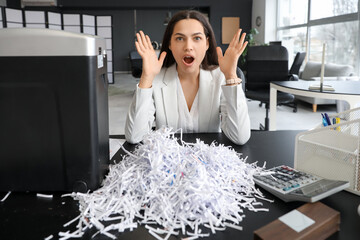 Shocked young secretary with shredded paper and shredder in office