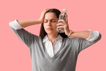 Young woman with bottle of water suffering from headache on pink background