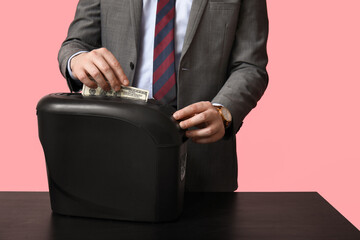 Young man in suit destroying dollar banknote in shredder on pink background, closeup