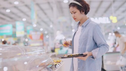 A woman is shopping for food in a store. She is holding a tray of food and looking at her phone