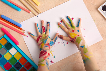 Child's hands in paint with paper sheet and different stationery for drawing on beige grunge...