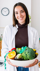 Vertical portrait of young nutritionist woman holding a bowl of natural and organic fruits and vegetables standing at office consultation. Healthy lifestyle and nutrition concept.