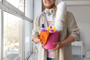 Young bearded man with bucket of cleaning supplies near window at home, closeup