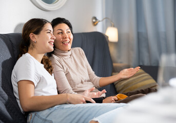 Two female friends sitting on couch, laughing and watching television.