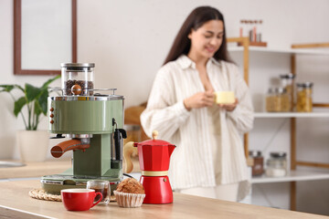 Pretty young woman with cup of hot espresso and coffee maker at home