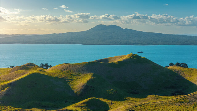 Aerial shot of a grassy volcanic crater on a coastal hillside with the ocean and distant Rangitoto in the background - Musick & Browns Island Motukorea