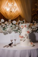 White wedding cake decorated with fresh roses. Three-tiered wedding cake decorated with fresh flowers and champagne glasses for the bride and groom on the table in the restaurant in the evening