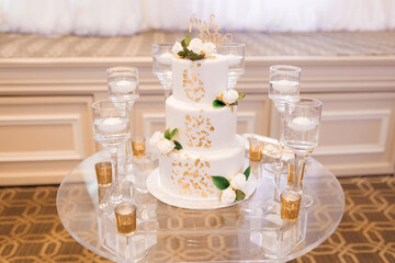 White three-tiered wedding cake with gold petals. Wedding cake with the letters Mr and Mrs on top decorated with white roses and gold details on a glass round table with candles around