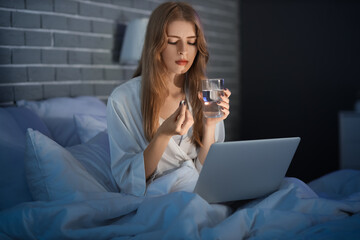 Young woman with laptop taking sleeping pill in bedroom at night