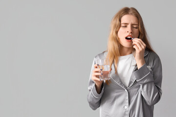 Tired young woman in pajamas taking sleeping pill on light background