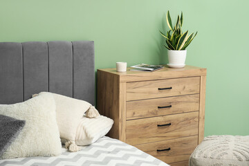 Wooden chest of drawers with houseplant and magazines near cozy bed in bedroom