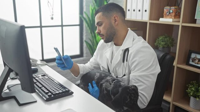 Young hispanic male veterinarian in a clinic using a mobile phone while holding a teckel dog on his lap inside an indoor workspace.